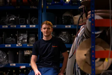 A young man sitting on a pallet in a warehouse surrounded by shelves of equipment and materials