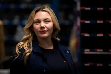 Woman with long blonde hair wearing a dark jacket in a warehouse setting