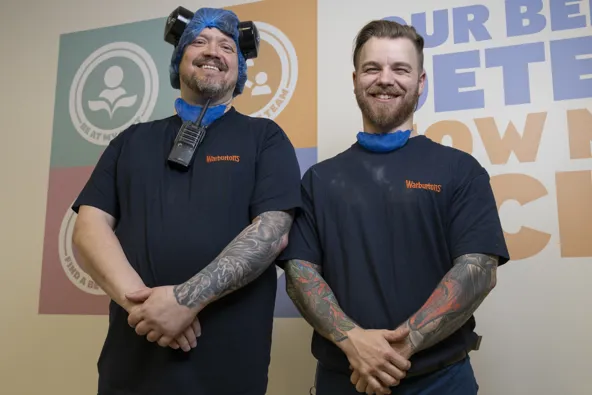 Two men wearing blue hairnets and black shirts smiling in front of a colorful wall with logos