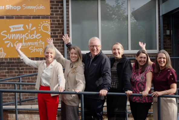 Group of six people smiling and waving in front of a community shop sign