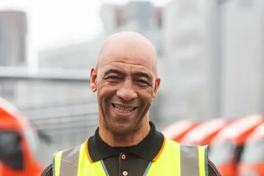 Man in a high-visibility vest smiling and holding a device, with trucks in the background