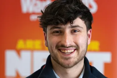 A smiling young man wearing a navy sweater and a lanyard with a logo