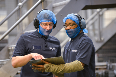 Two workers in protective gear discussing information on a tablet in a manufacturing setting